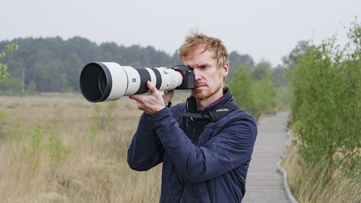 Man holding the Sony A7R VI up to his eye with the FE 400-800mm F6.3-8 G OSS supertelephoto zoom lens attached, he's standing on a boardwalk in a nature reserve