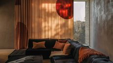 The corner of a modern living room with a floor-to-ceiling window with a peach colored curtain with an abstract red print. A large dark blue corner sofa with warm rust-colored cushions and throws. 
