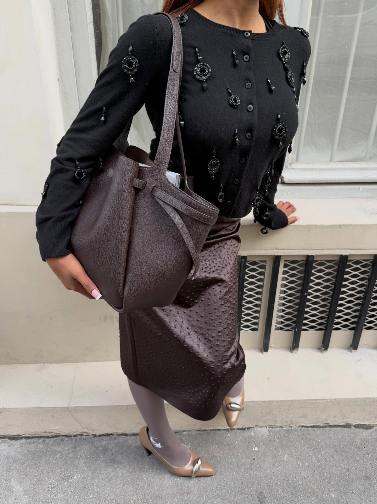 An image of an influencer in a black beaded cardigan, brown ostrich leather skirt, mauve tights and beige Tory Burch pierced shoes holding a big leather tote.