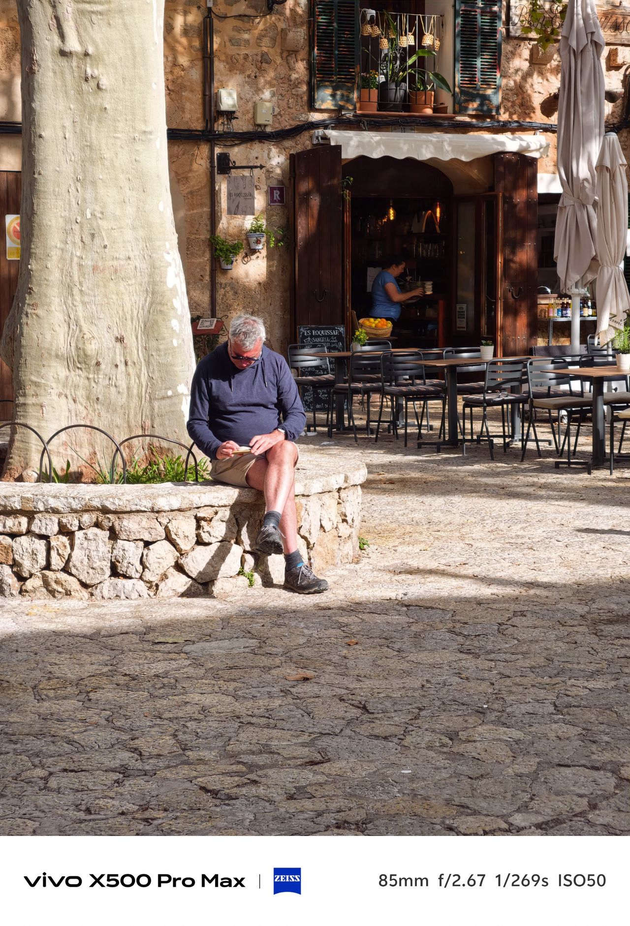 Man sitting beneath a large tree beside an outdoor cafe in a sunlit stone village square.