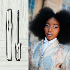 A woman wearing white eyeshadow with long lashes and her curls out in an afro. She is also wearing a pastel blazer and button-up shirt. She is next to a tube of mascara. 