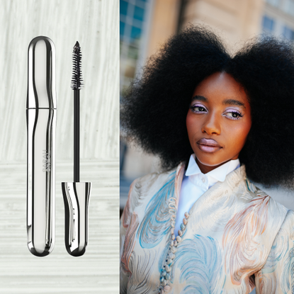 A woman wearing white eyeshadow with long lashes and her curls out in an afro. She is also wearing a pastel blazer and button-up shirt. She is next to a tube of mascara.