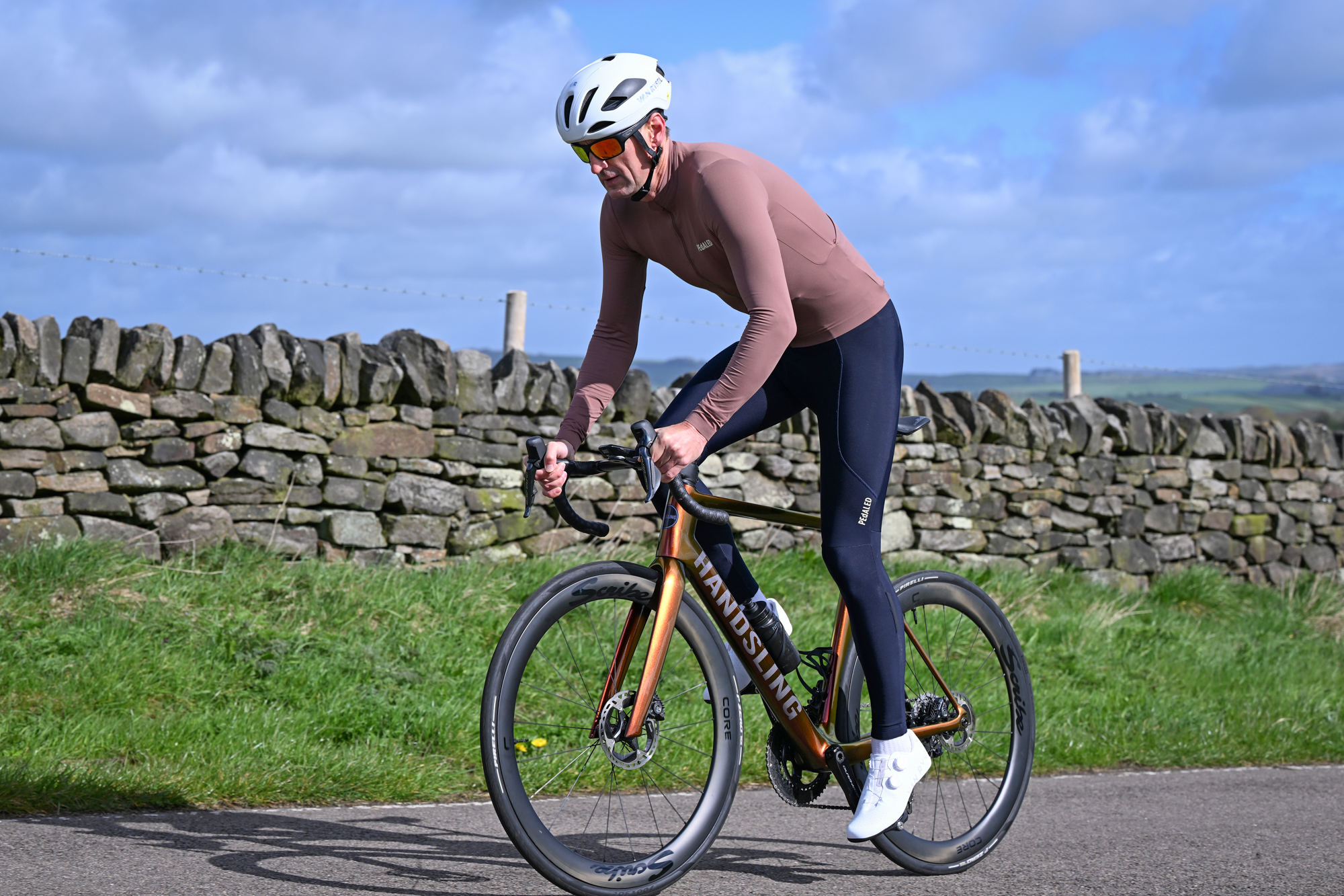 Man riding an orange bike out of the saddle on a country lane, wearing dark blue tights, purple jersey, white helmet and white shoes