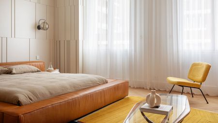 neutral bedroom with tan leather bedframe, bright yellow chair, and bright yellow rug