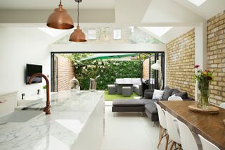 a modern, open plan kitchen extension, with an exposed brick wall to the right, bifold doors to the garden, a large marble island with bronze hanging lights, and a wooden dining table and dark grey sofa to the right