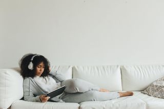 Eine Frau chillt auf dem Sofa, mit Kopfhörern auf den Ohren und einem Tablet in der Hand
