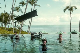 A series of film practitioners intent on filming a scene is fully immersed in an infinity pools while holding shooting gear against a tropical background.