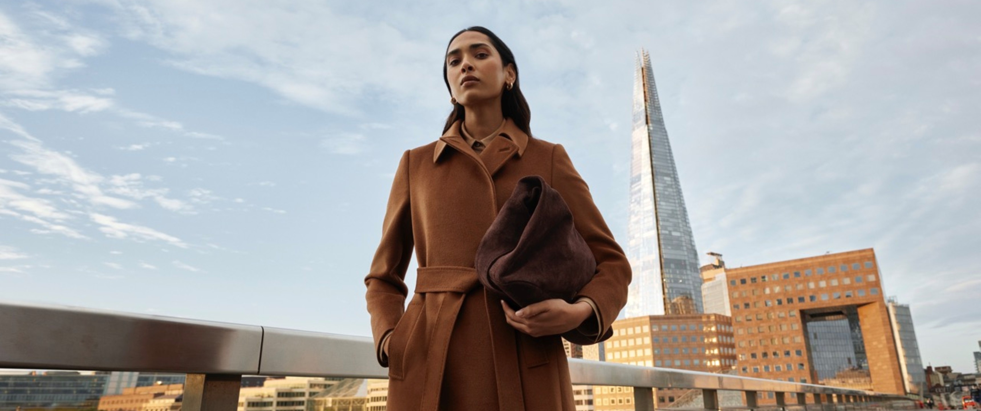 Model wearing a light brown wool coat holding a brown suede bag sold at Hobbs in front of the Shard