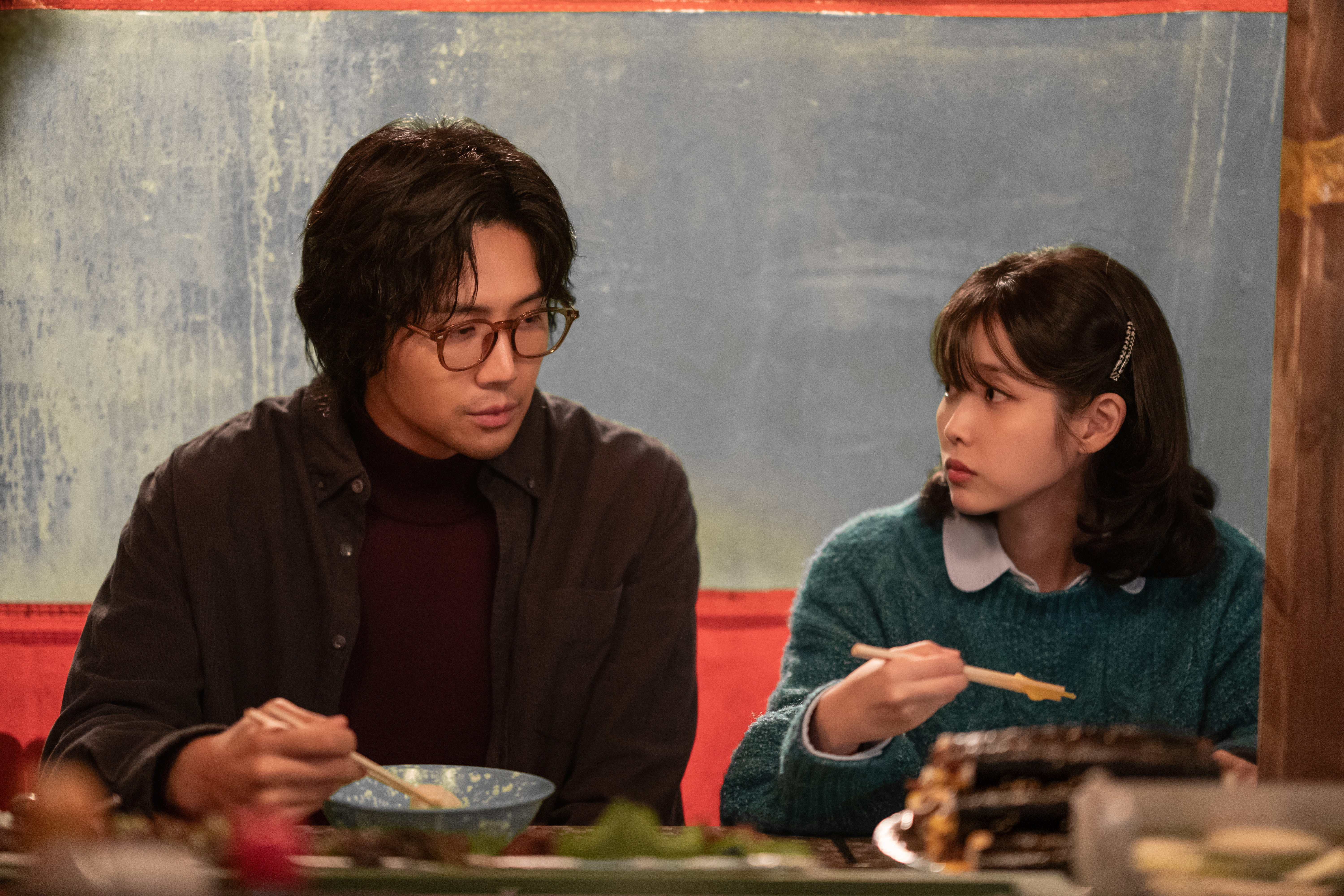A man with long hair and glasses (Kim Seon-ho as Park Chung-seop) and a woman in a green sweater (IU as Yang Geum-myeong) eat a meal in a Korean pocha, in a still from Netflix's 'When Life Gives You Tangerines.'