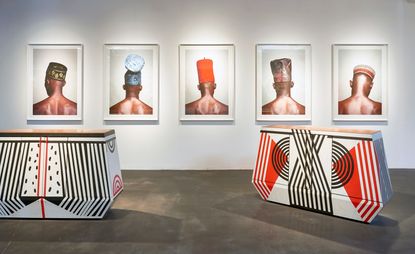 View of two black, white and orange patterned cabinets and framed portraits on the wall of men wearing different hats shot from the back. The space features white walls and grey floors