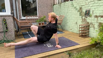 Harry Bullmore performs a kick through move on a yoga mat in his garden. He is concentrating as he exercises low to the ground, with one hand behind him for balance, one hand resting near his temple, one leg bent with his foot resting on the floor, and one leg held straight and slightly above the ground. 