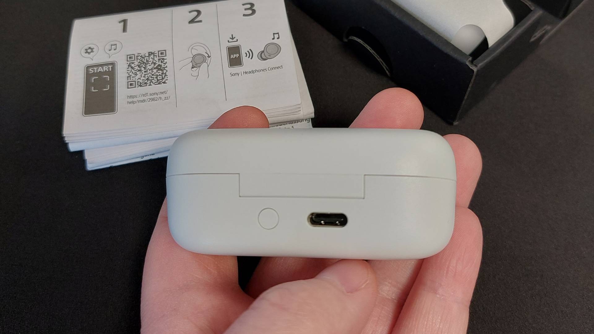 The Sony WF-C510 earbuds on a black desk mat. They are seen here inside their sleek charging case. This image focusses on the back, showing the pairing button and USB-C charging port.