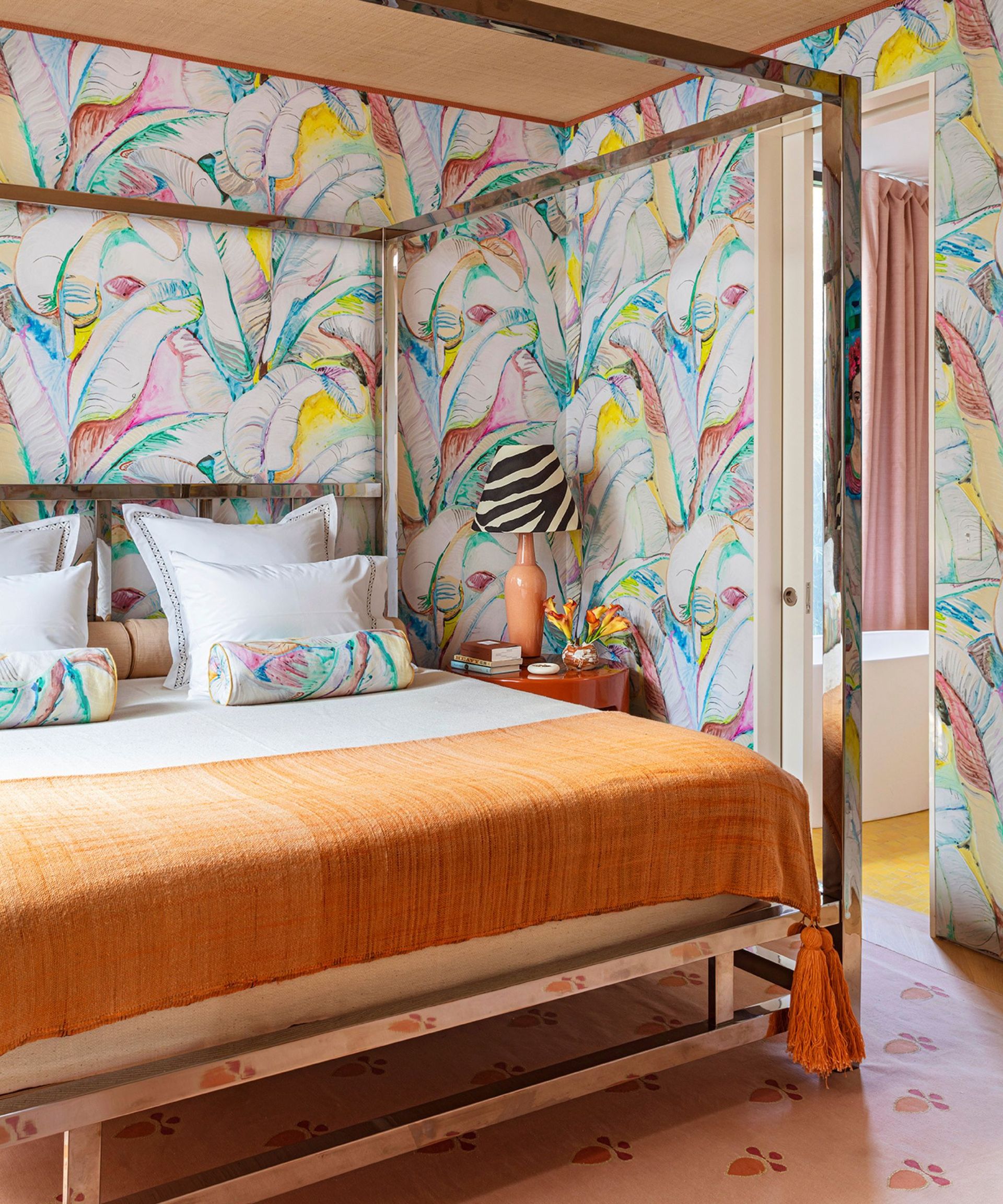Colourful bedroom with printed wallpaper, pink and orange carpet and bed with orange throw and four poster frame