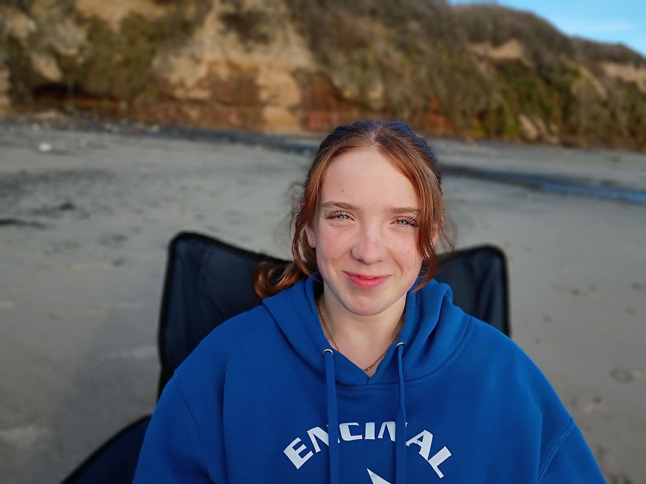 person with dark hair smiling wearing a blue hoody sitting on a black beach chair in front of a cliff on the sand in late-afternoon sun