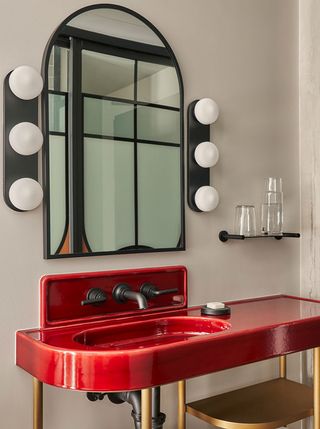 The creatively designed bathroom of a hotel features dressing room-inspired mirror and lights and a red and gold polished sink with an industrial feel.