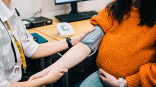A pregnant woman in an orange sweater gets her blood pressure monitored by someone wearing a white shirt and a stethoscope.