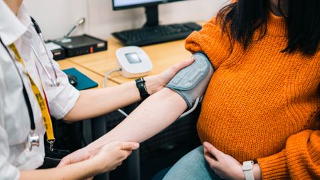 A pregnant woman in an orange sweater gets her blood pressure monitored by someone wearing a white shirt and a stethoscope.