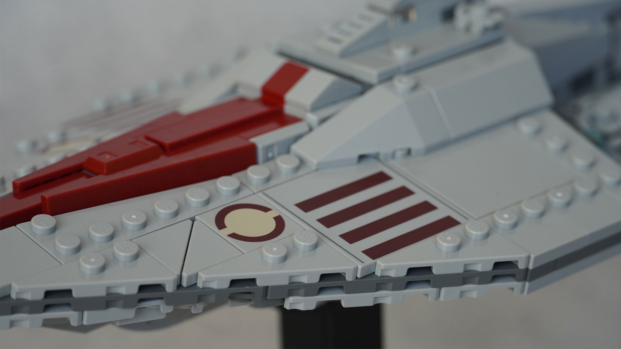 A miniature-sized long grey ship made of bricks with a red stripe running down the middle is placed on a black stand in front of a grey background.