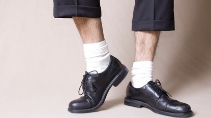 A man's bare legs show between the hems of his trousers and the tops of his white socks and block shoes. 