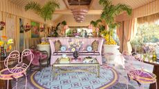 Yellow cabana-striped tiki room featuring floral pastel furniture and rugs, accented by oversized leafy plants and a feathered glass chandelier