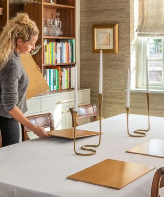 A table being set up for Christmas dinner with a white tablecloth, gold rectangular placemats, and two gold double taper holders at the center