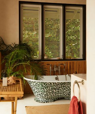 Taupe bathroom with bright coral towel, white shutters in black frame, and tropical theme with large palm plant, bamboo paneling an leopard print tub