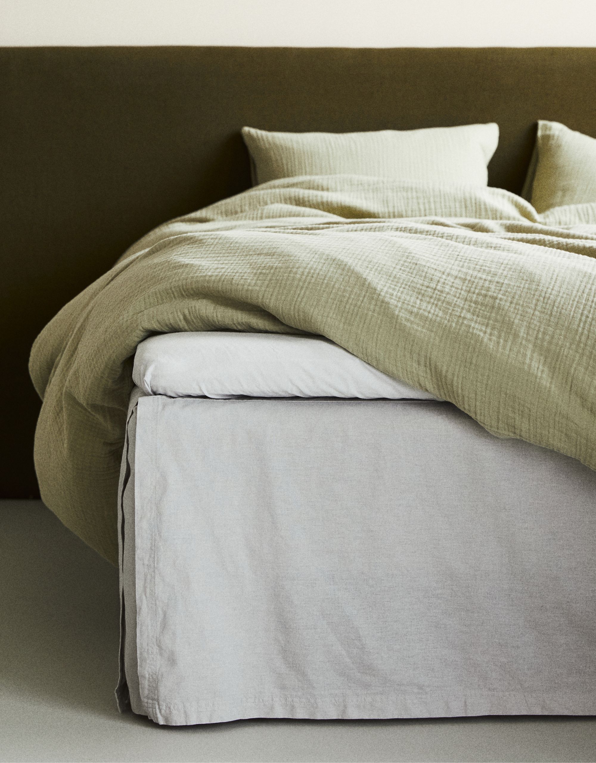 The side of a bed with off white-gray sheets and valance and a light green muslin duvet cover draped across the bed at an angle with a dark green velvet headboard