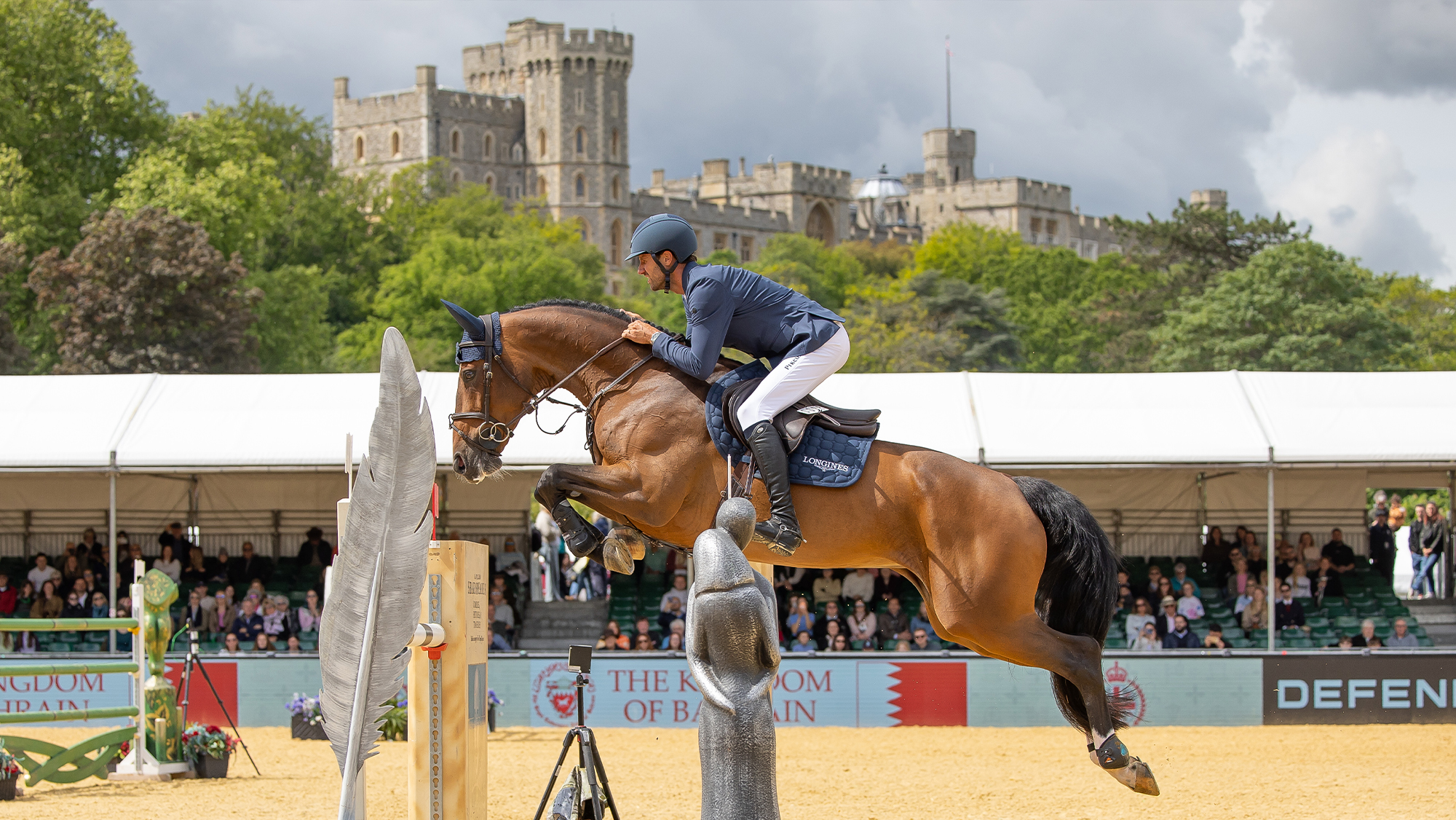 Male showjumper jumping bay horse in main arena at Royal Windsor Horse Show with Windsor Castle in the background