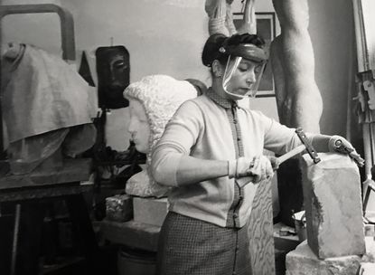 Naomi Feinberg in her workshop, holding a sculpting tool on a brick