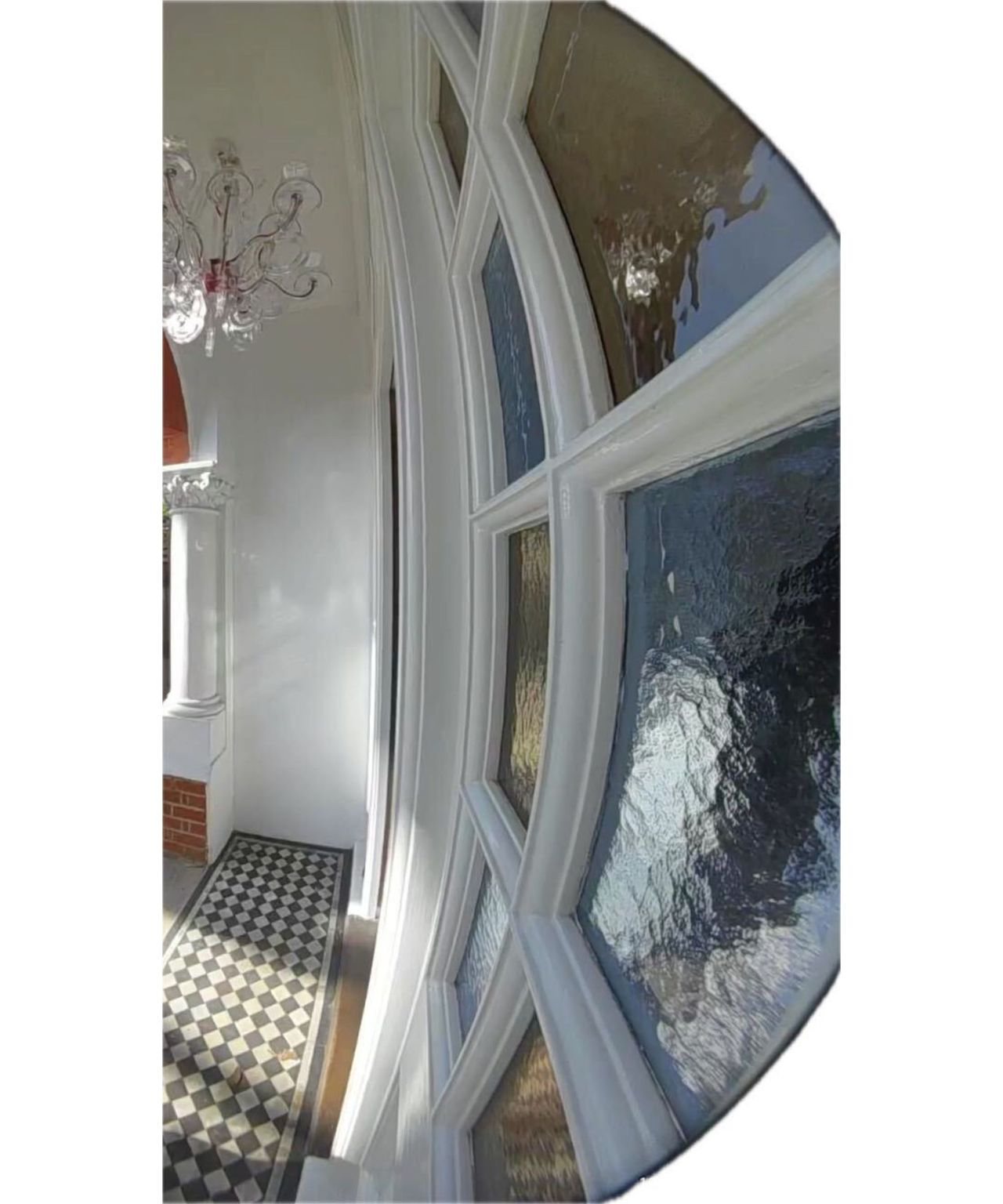 The view of a black and white tiled porch from Ring Battery Doorbell Plus. To the right are stained glass panels, and above is an acrylic light with pink wiring.