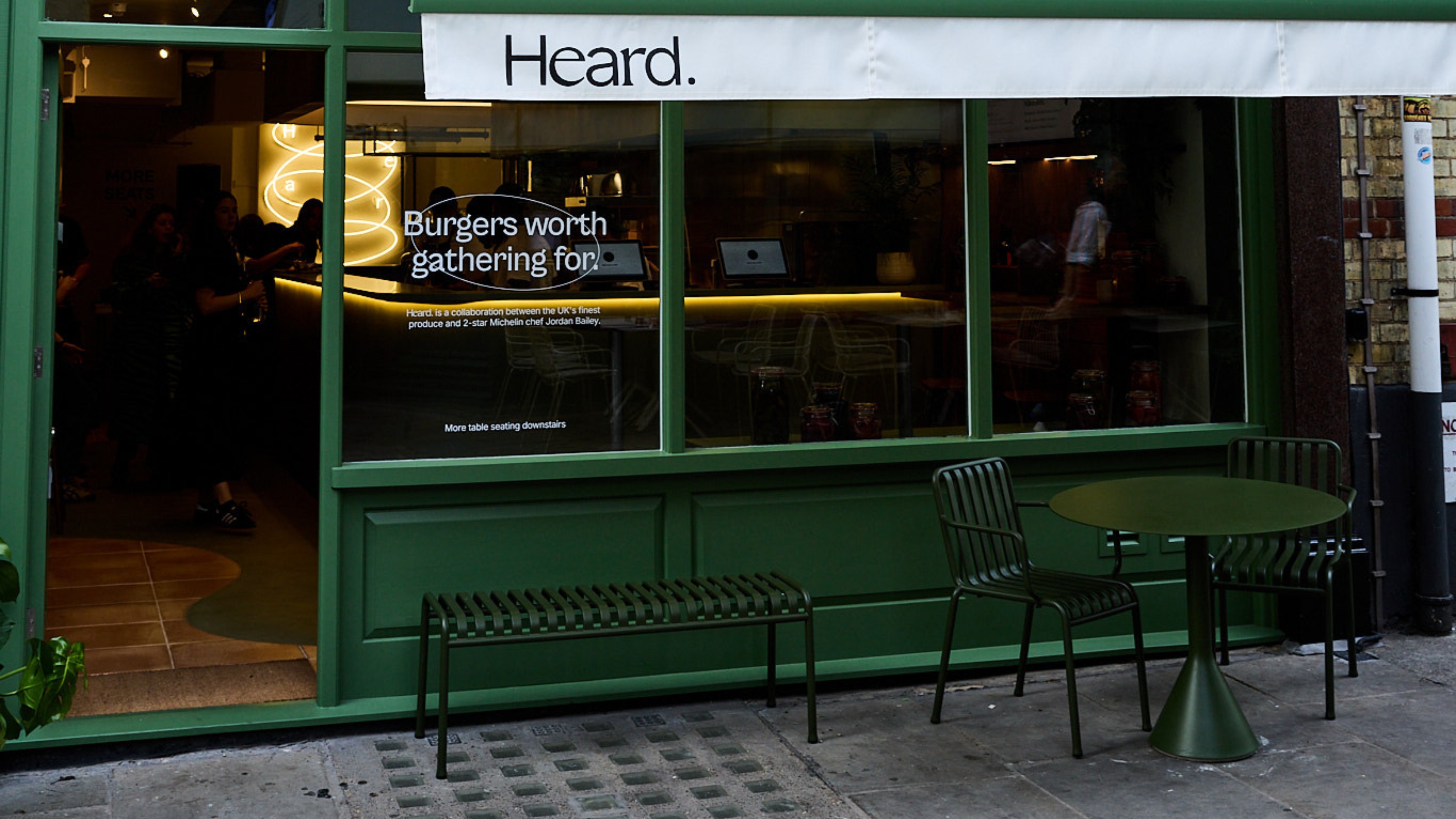 The green-painted exterior of a high-end burger join features lacquered emerald green furniture in iron and lets passersby peek inside its atmospheric, sculpturally designed eatery with neon-lit signs.