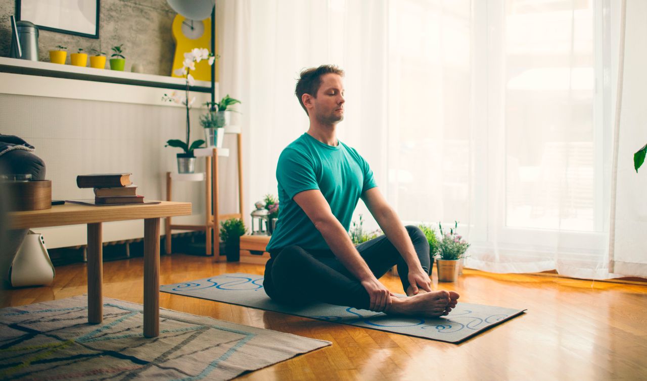 Man sits on a yoga mat in a living room with the soles of his feet together and his knees out to the sides. He wears grey yoga pants and a green T-shirt