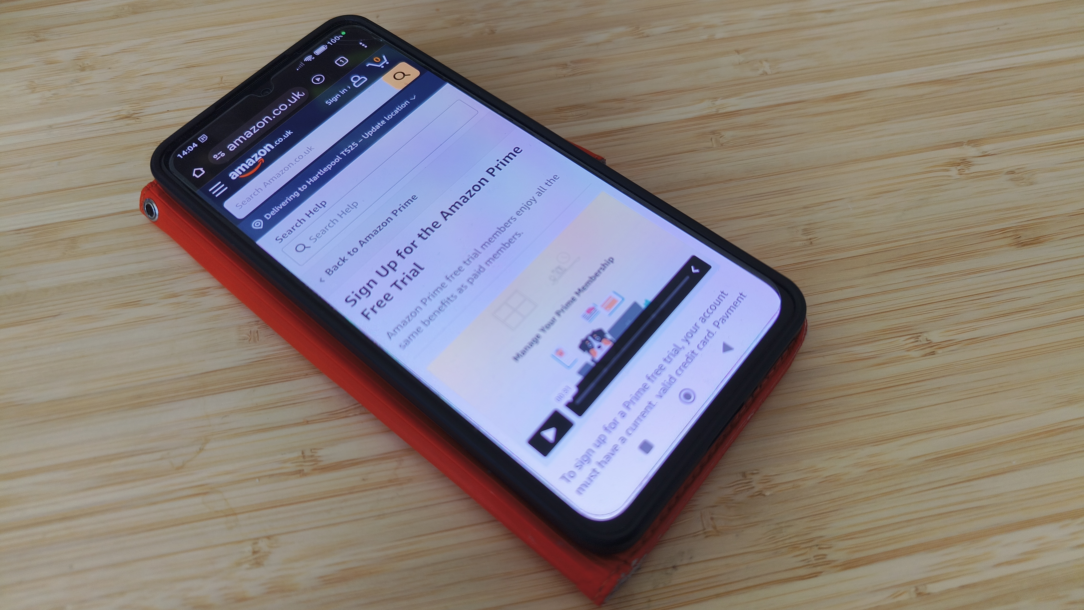 A smartphone with an orange case displaying the Amazon Prime sign-up page on its screen, resting on a wooden surface.