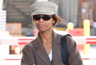 Zoe Saldana is seen at John F. Kennedy International Airport on October 14, 2024 in New York City wearing a hat, brown coat, black cardigan, jeans, and black Saint Laurent loafers. She is carrying a Saint Laurent shoulder bag.