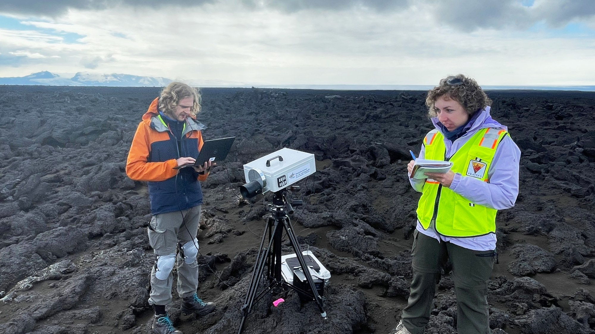 two people stand on a black lava field with a white insrument on a black tripod