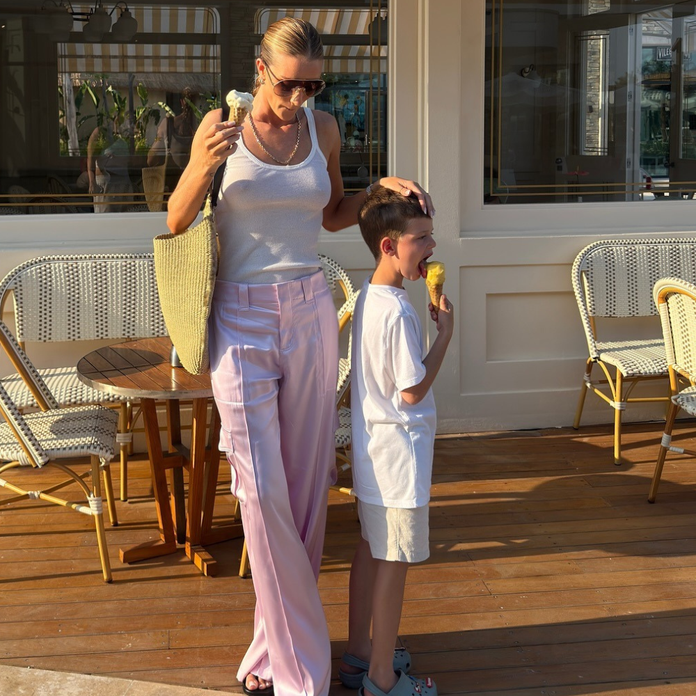 Fashion person Rosie Huntington-Whiteley wears the pale pink trouser trend. She pairs her trousers with a white tank top, khaki raffia tote and thong sandals on holiday. She is looking down, holding her son's head in front of a old-fashioned ice-cream shop in a recent image from her Instagram. 