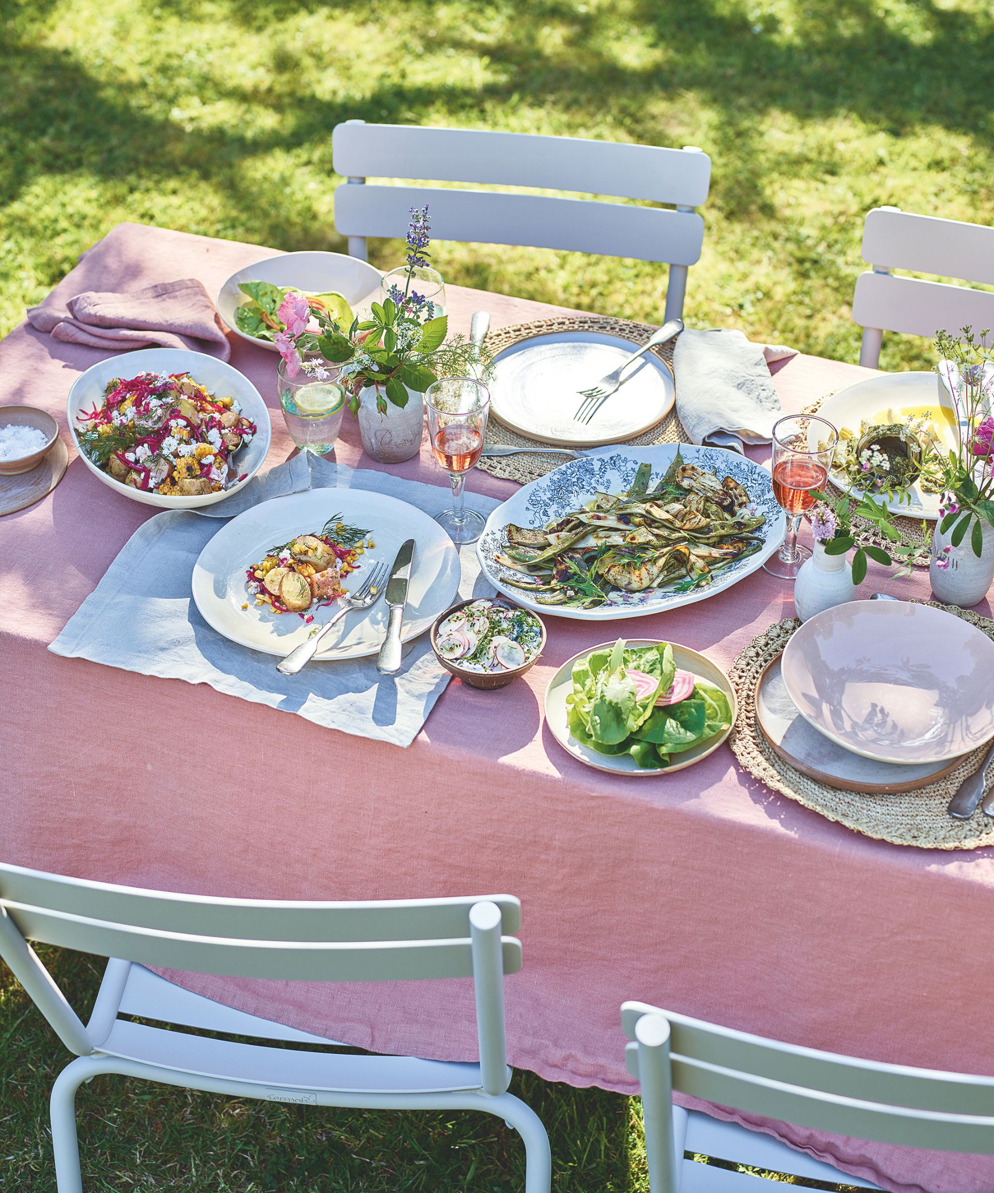 Outdoor dining table with metal bistro chairs, pink linen tablecloth, plates of grilled vegetables and salads, rattan placemats, white plates, wine glasses, and bud vases filled with fresh stems.