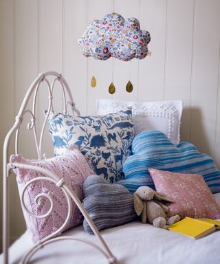 A baby girl nursery idea with a daybed, selection of textured cushions and a rain cloud mobile in whites and pastels.