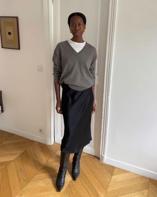 style influencer Sylvie Mus wearing a winter outfit with a gray v-neck sweater layered over a white tee, black satin skirt, and black pointed-toe knee-high boots