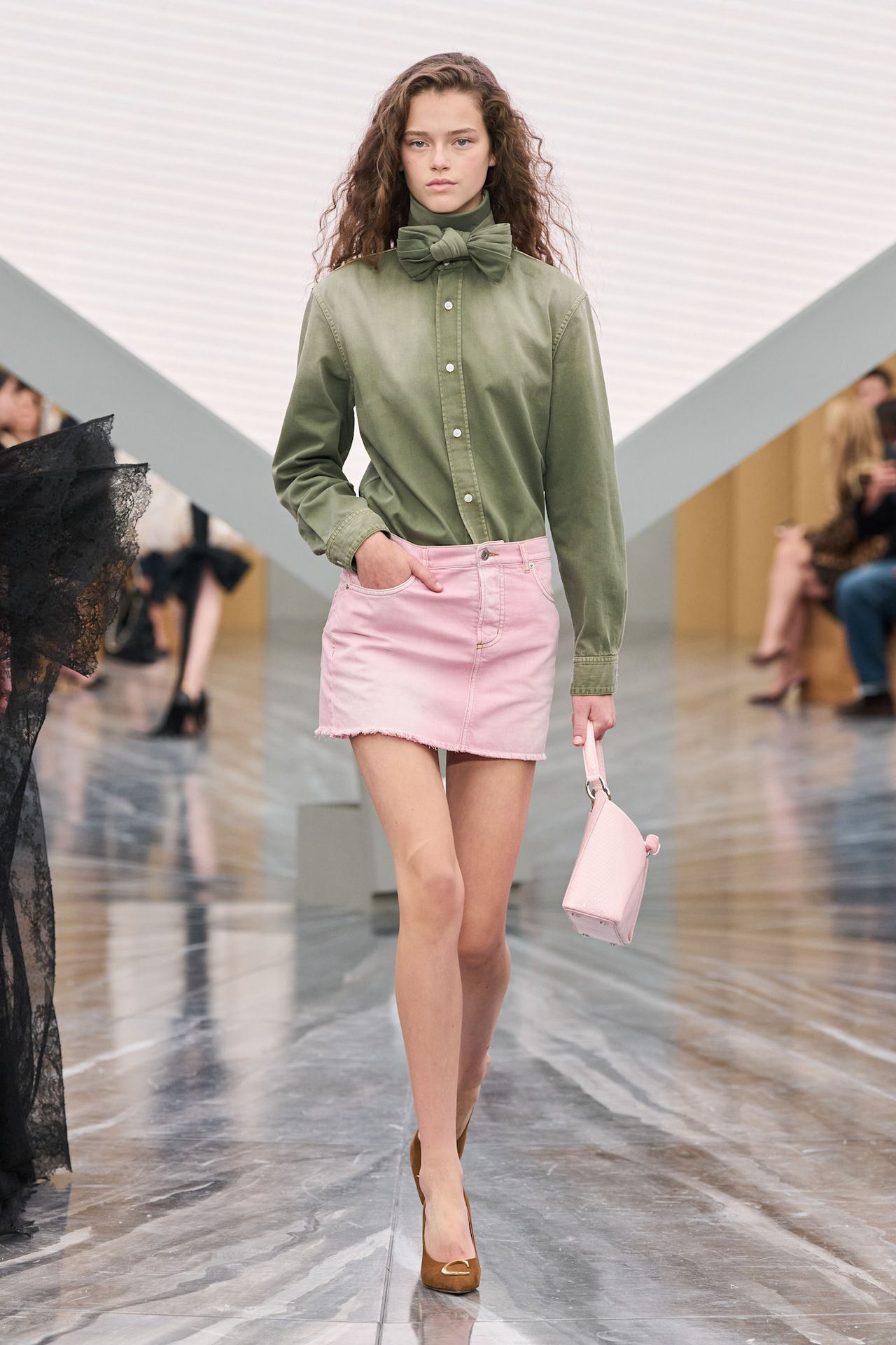 A Dior model wearing a sage green button-down shirt with a bow at the collar, a pale pink denim miniskirt, a pale pink handbag, and tan pointed pumps.