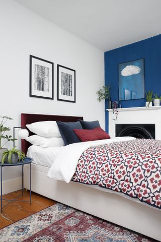 Bedroom with white walls, one blue feature wall, burgundy velvet headboard and red and blue pattern bedding, red Persian-style rug and framed artwork