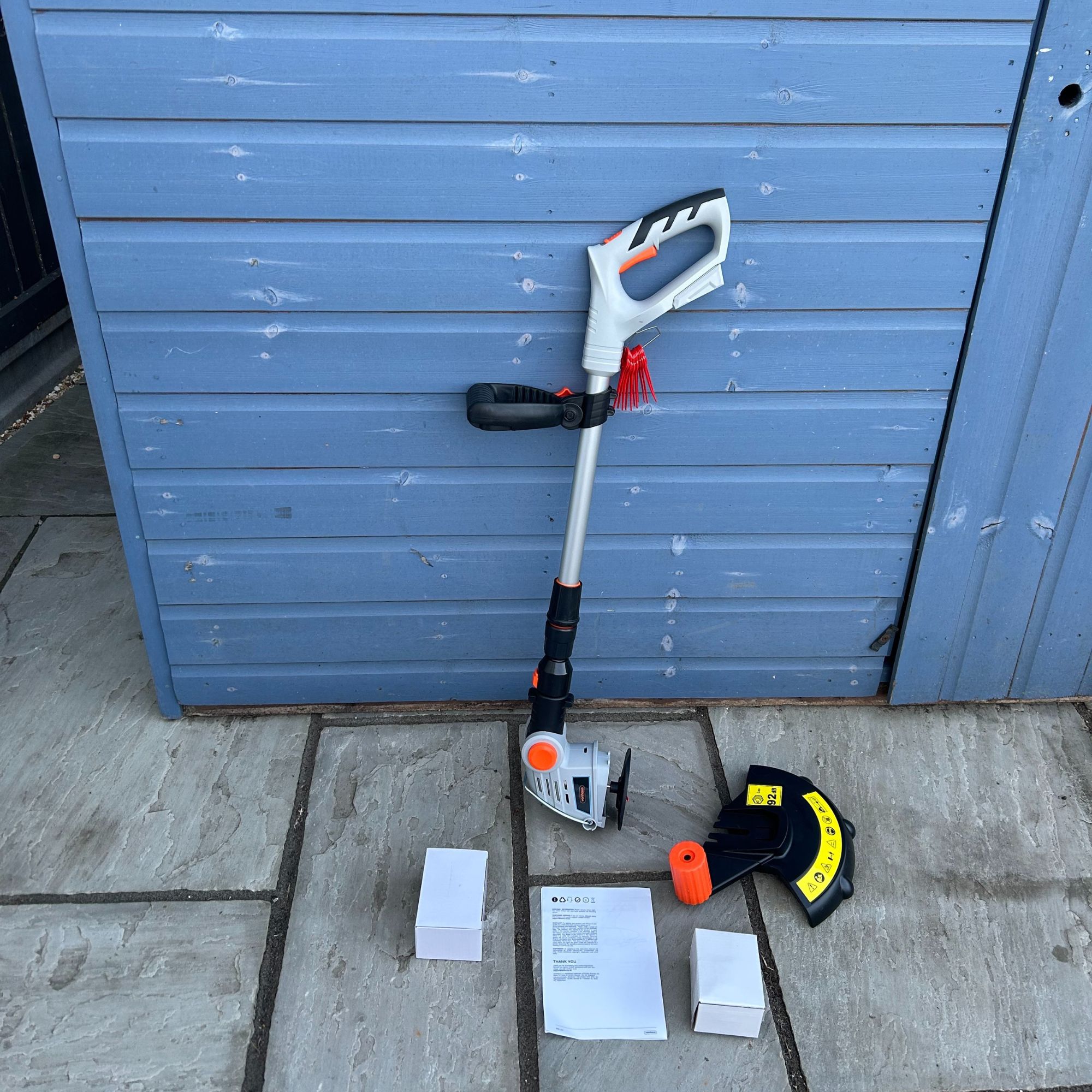 VonHaus G-Series Cordless Grass Trimmer box contents on floor against blue shed