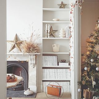 A white living room with a marble fireplace decorated for Christmas with a Christmas tree in the adjoining room decorated in white and gold