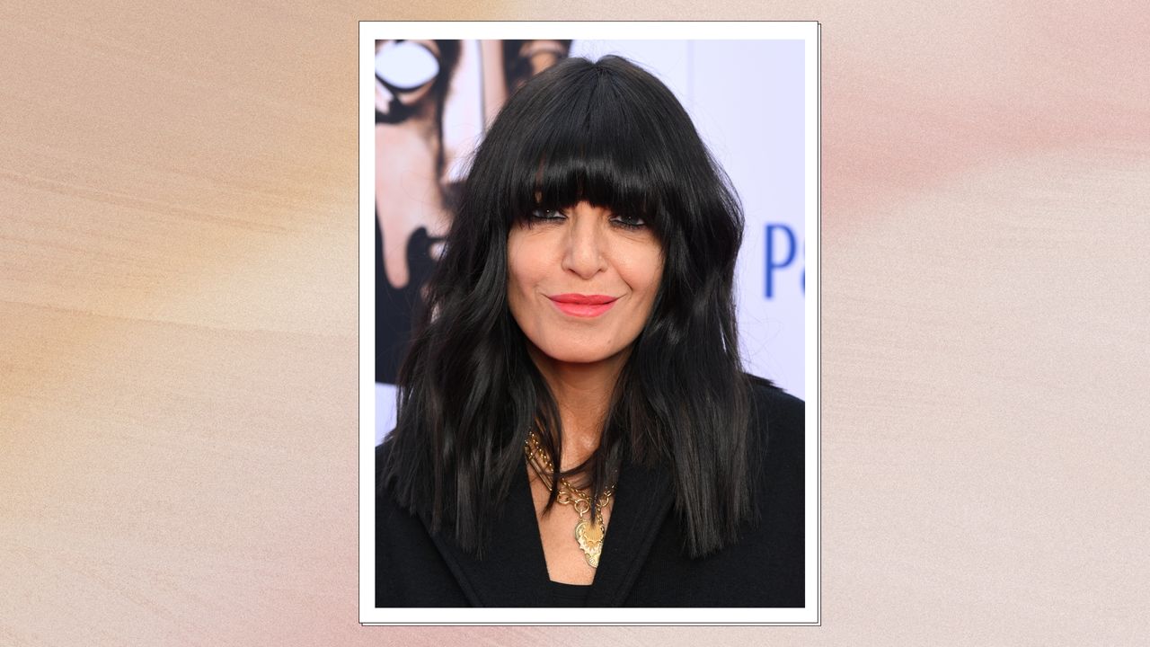 Image of Claudia Winkleman with black wavy hair with full fringe, smiling and wearing a coral lipstick, with a pink watercolour background