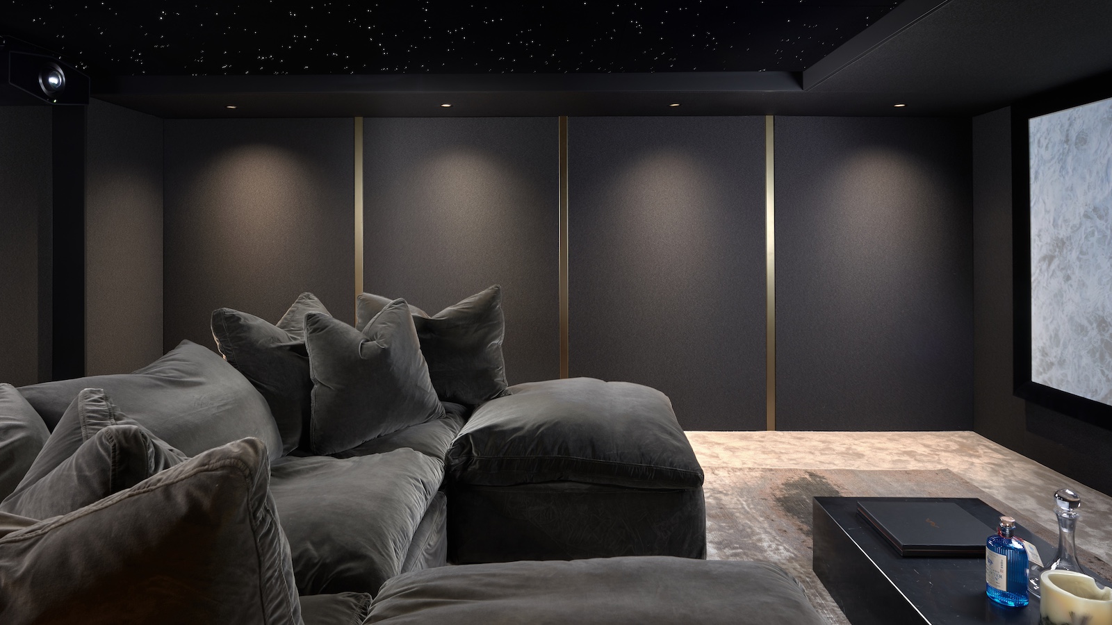 A photograph of a luxury, custom-installed home cinema with a huge projector screen, lush u-shaped sofa, grey walls and lamps