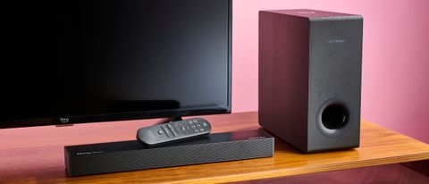 Ultimea Nova S50 soundbar with remote resting on top of it. There is an Amazon Fire TV behind the soundbar, and the Nova S50 subwoofer is positioned to the right of the TV