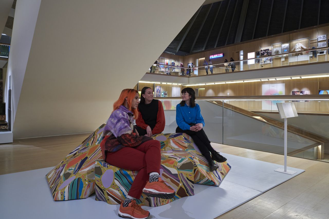 A soft seating area in a museum has a scattered, rock-like sofa with a colorful, mineral-like surface as its focal point, and three young women sitting on it.