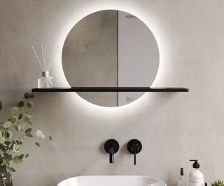 round framelessbacklit mirror with black shelf on white wall with black taps underneath, white sink green plant and soap dispenser