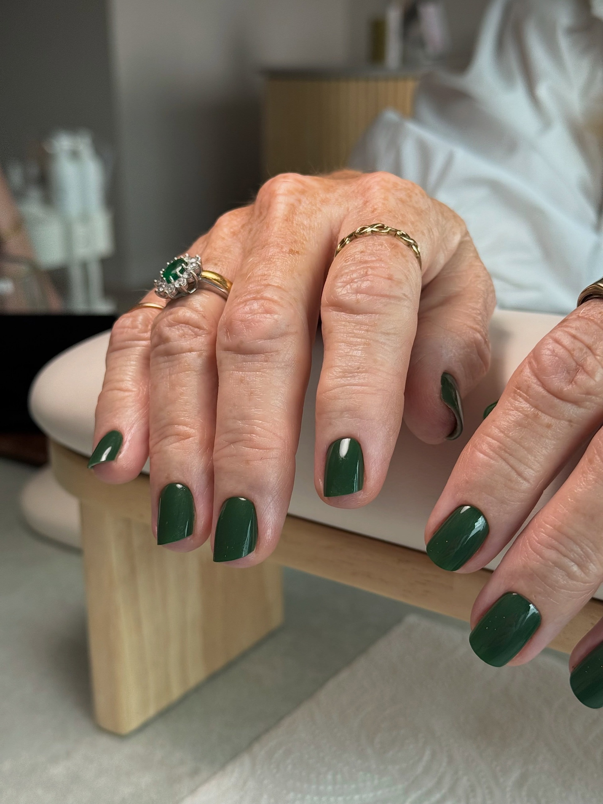 A pair of hands with glossy dark green nails and gold rings, including a large green gemstone ring