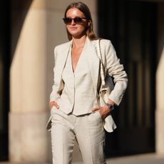 Diane Batoukina wears a light beige linen suit consisting of a single-breasted blazer with notched lapels and matching trousers. The blazer features a tailored fit with buttoned cuffs. Underneath, a matching waistcoat with a V-neckline and button fastening is visible. Sunglasses with dark lenses and a tortoiseshell frame are worn. A white leather shoulder bag with a gold chain strap is carried. A metallic watch adorns the wrist. Hair is styled straight and parted in the middle, during a street style fashion photo session, on May 14, 2025 in Paris, France